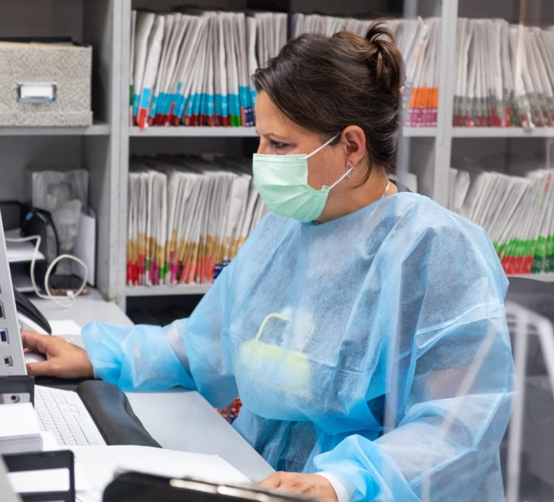 receptionist at dental office wearing mask and gown for protection during the Covid-19 pandemic. She is working at the computer with patient files in the background.