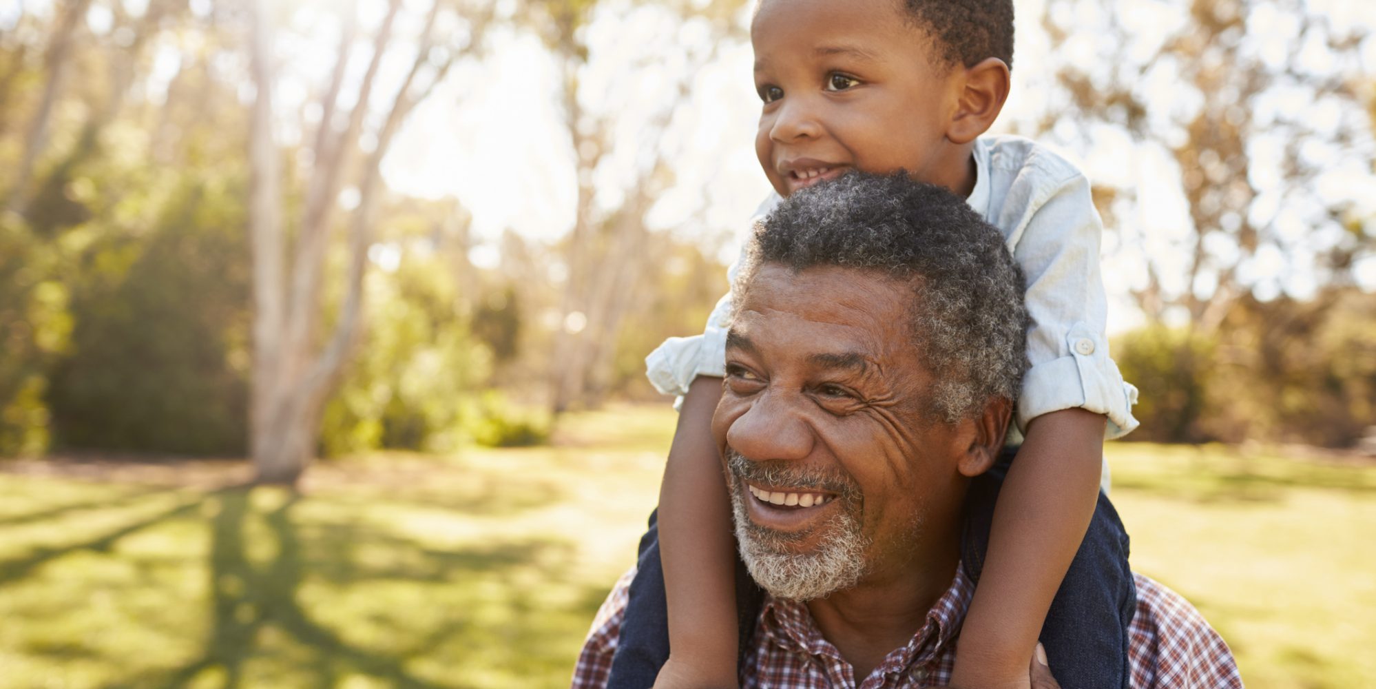 Image of grandfather carrying grandson on shoulders through park - dependent